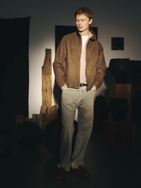 Adult male wearing a brown jacket and light-wash jeans stands indoors in a fashion editorial. He's illuminated by directional light.