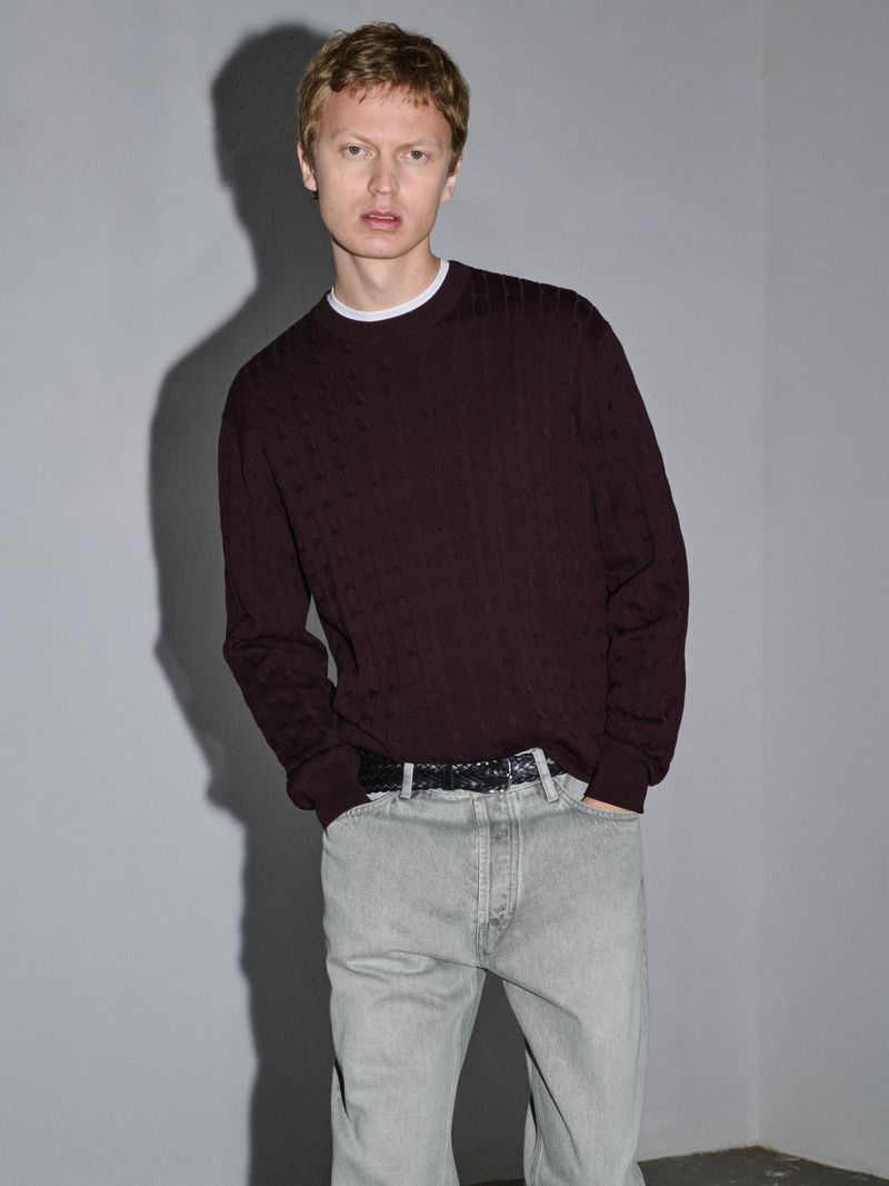 A male model wearing a dark burgundy sweater and light wash jeans standing against a plain grey studio wall.