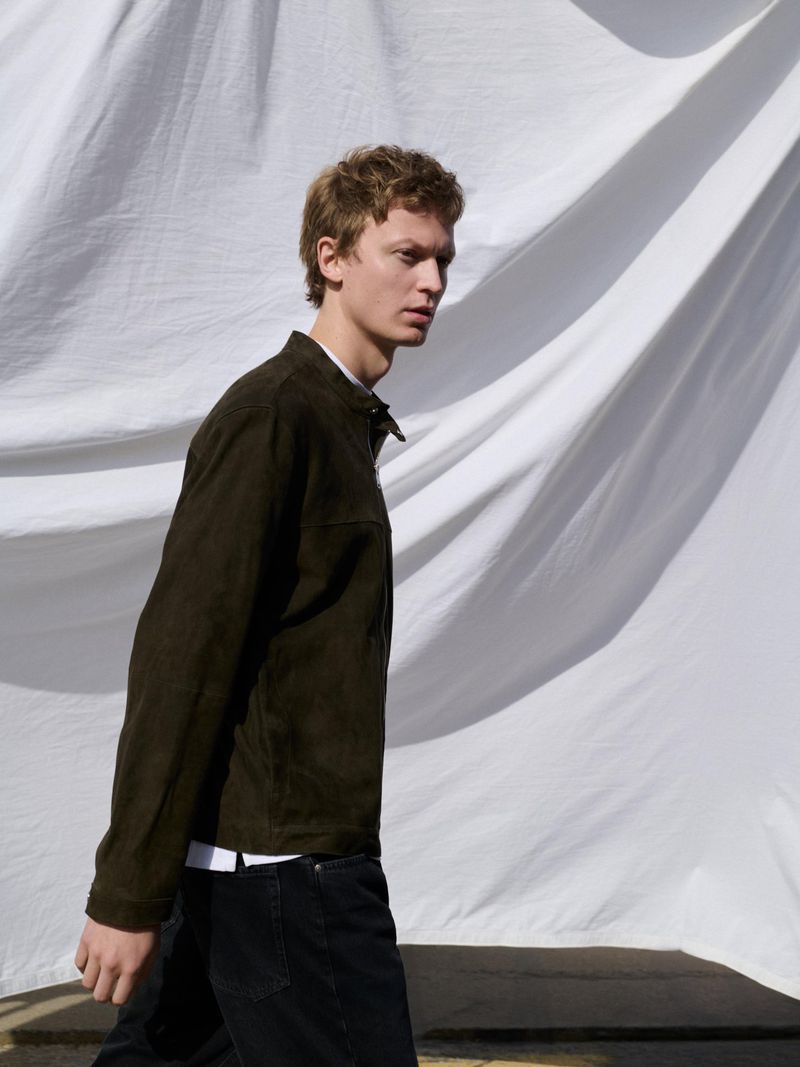 A young man wearing a dark jacket and jeans walks in front of a white fabric backdrop, shot in natural light with a side view.