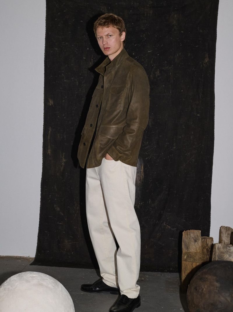 A fair-skinned man poses indoors, wearing an olive jacket and cream pants, showcasing modern fashion against a neutral background.