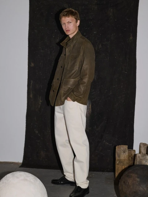 A fair-skinned man poses indoors, wearing an olive jacket and cream pants, showcasing modern fashion against a neutral background.