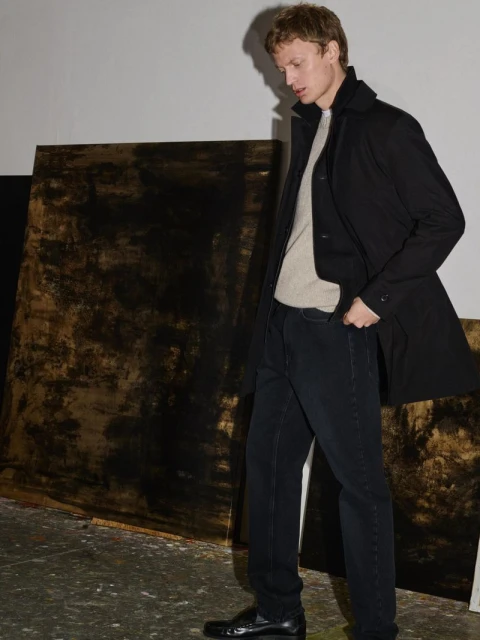 Indoor studio shot of a male fashion model in a black coat, light sweater, and dark jeans, standing near abstract painted backdrops.