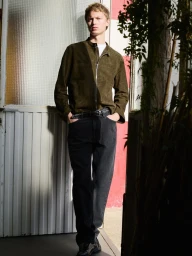 Full-length portrait of a male model wearing an olive green jacket and dark jeans standing against a doorway.