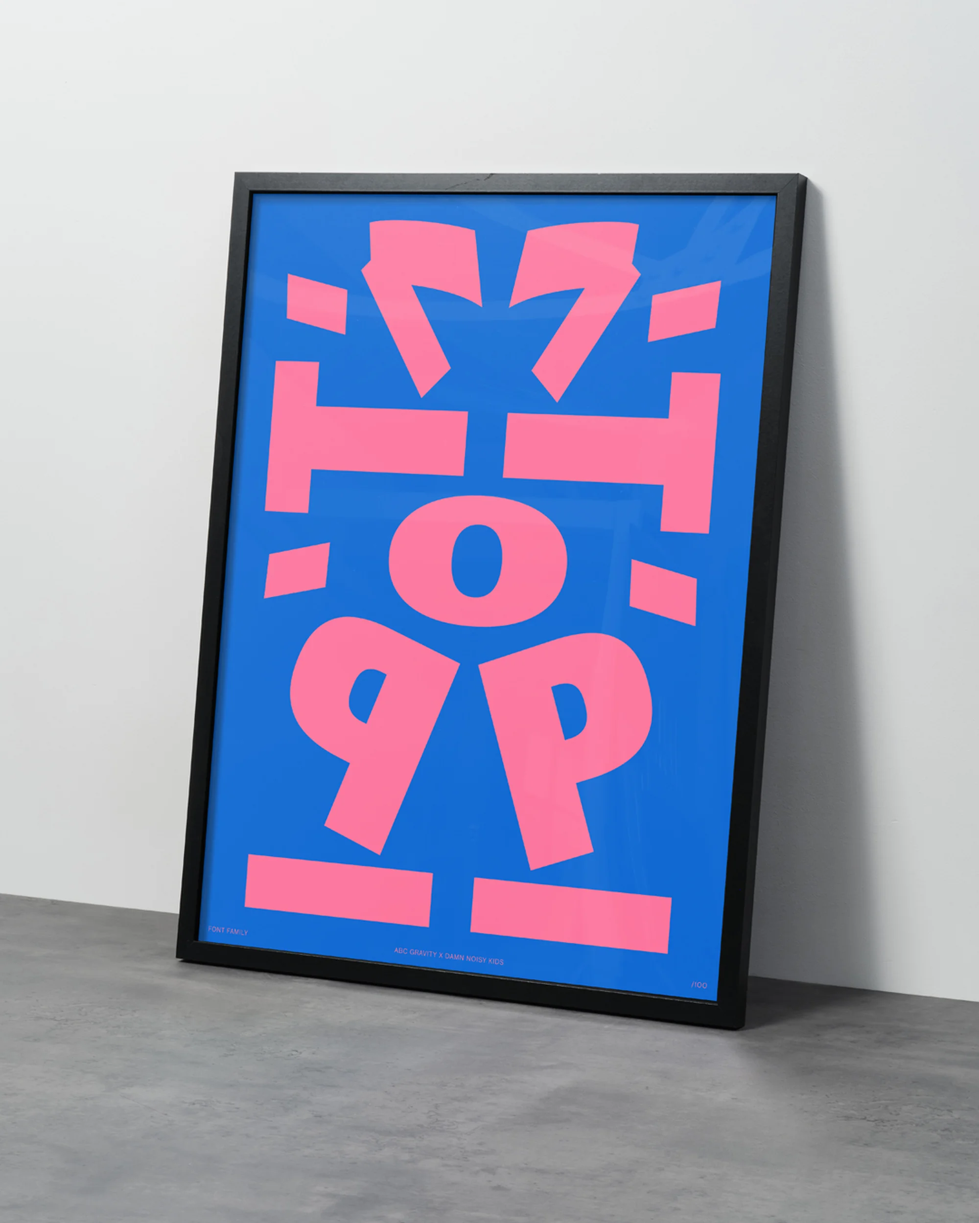 Framed graphic art print with pink abstract letters on a blue background. The artwork leans against a white wall atop a gray floor.