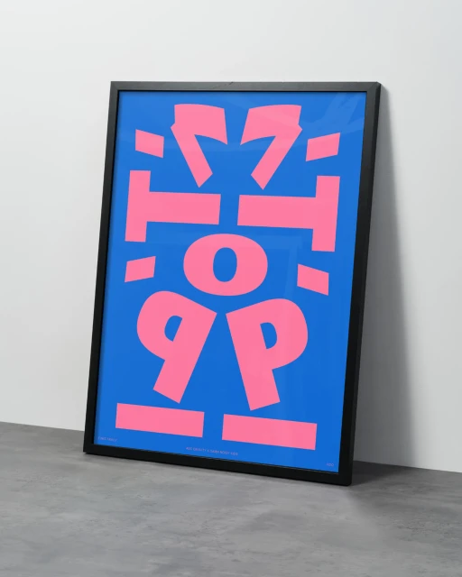 Framed graphic art print with pink abstract letters on a blue background. The artwork leans against a white wall atop a gray floor.