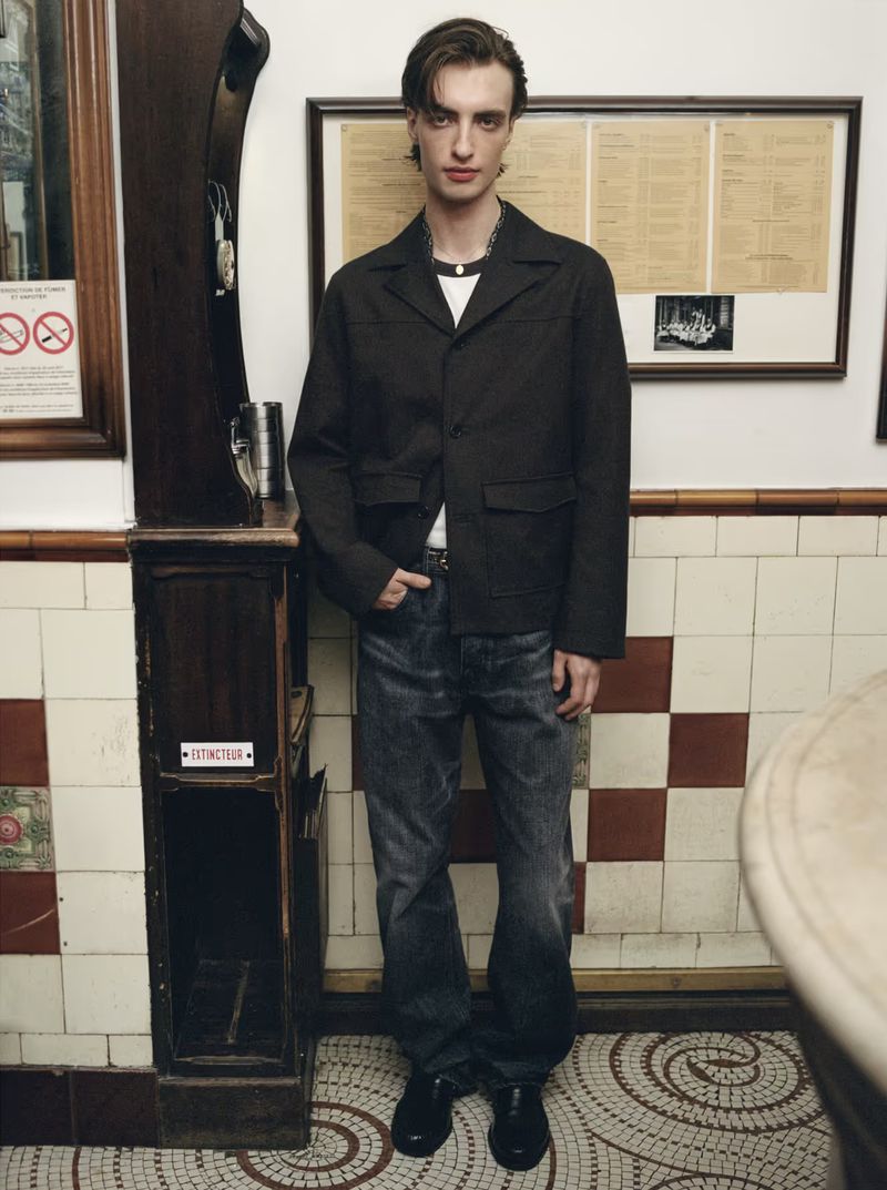 Man poses in an indoor setting wearing a dark jacket, jeans, and dress shoes, standing against a tile wall and wood paneling.