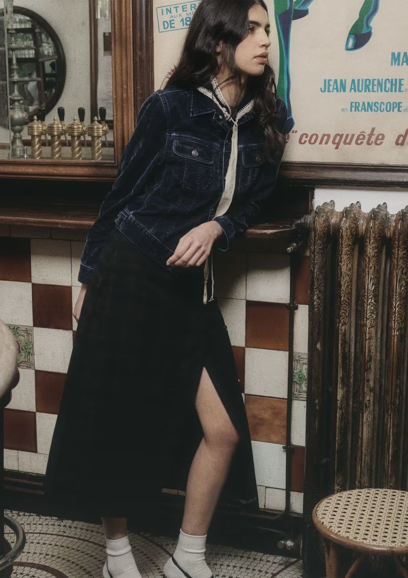 Indoor shot of a woman in a blue denim jacket and black skirt, leaning against a poster, showcasing fashion in a cafe.
