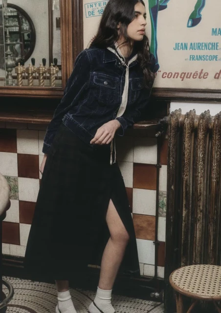 Indoor shot of a woman in a blue denim jacket and black skirt, leaning against a poster, showcasing fashion in a cafe.