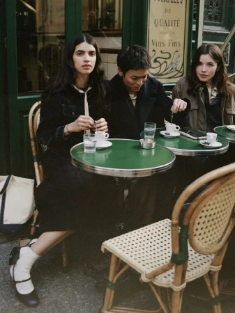 Three people sit at an outdoor cafe table, enjoying coffee and conversation in a European city setting with green tables and wicker chairs.
