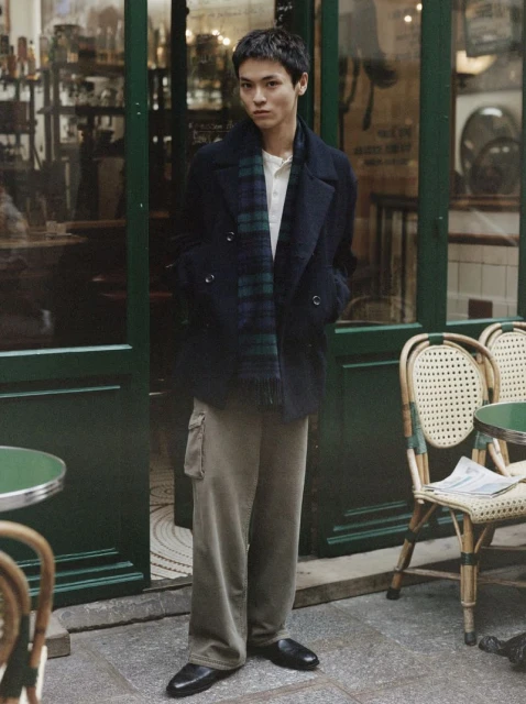 Male model in a pea coat and plaid scarf poses in front of a green door and chairs at an outdoor cafe in an urban setting.