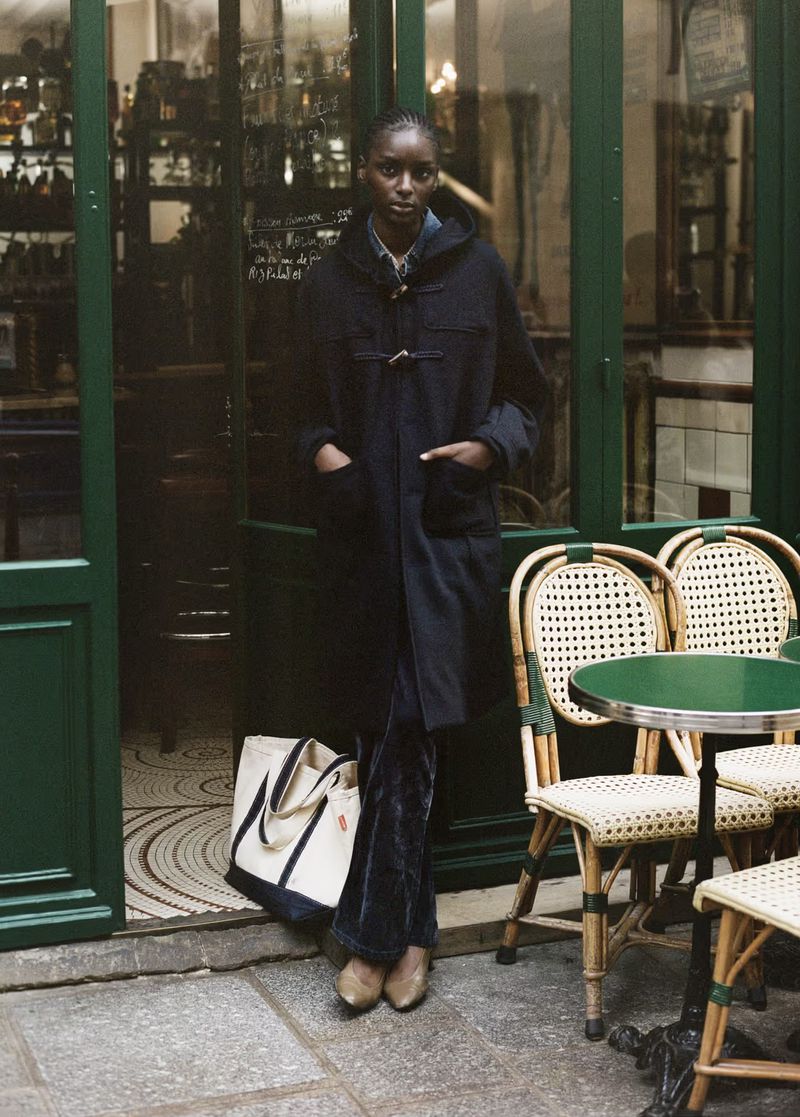 An African woman in a black coat and crushed velvet pants stands near a Parisian cafe entrance, a cream tote bag at her feet.