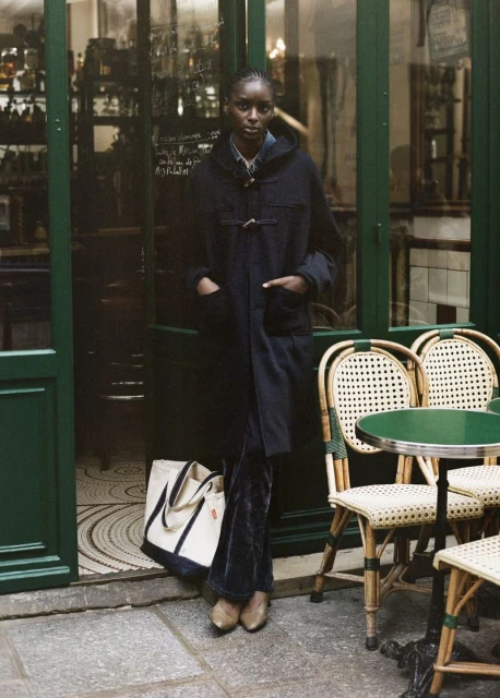 An African woman in a black coat and crushed velvet pants stands near a Parisian cafe entrance, a cream tote bag at her feet.
