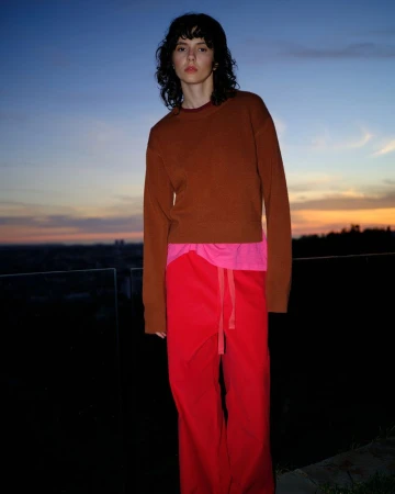 Young woman posing in earth-toned sweater and red pants against a twilight sky, standing outdoors in a portrait-style medium shot.