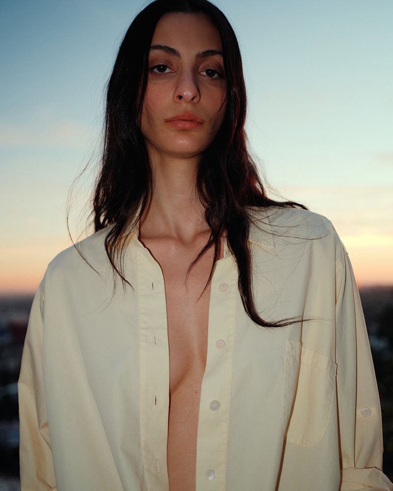 Close-up of a young adult woman with long dark hair wearing an unbuttoned yellow shirt against a blurry urban backdrop at dusk.