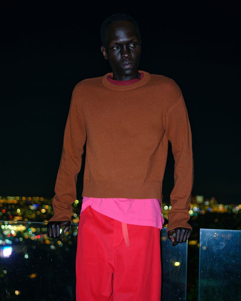 An androgynous person with a dark complexion stands outdoors at night, wearing an orange sweater and bright pink pants, city lights behind them.