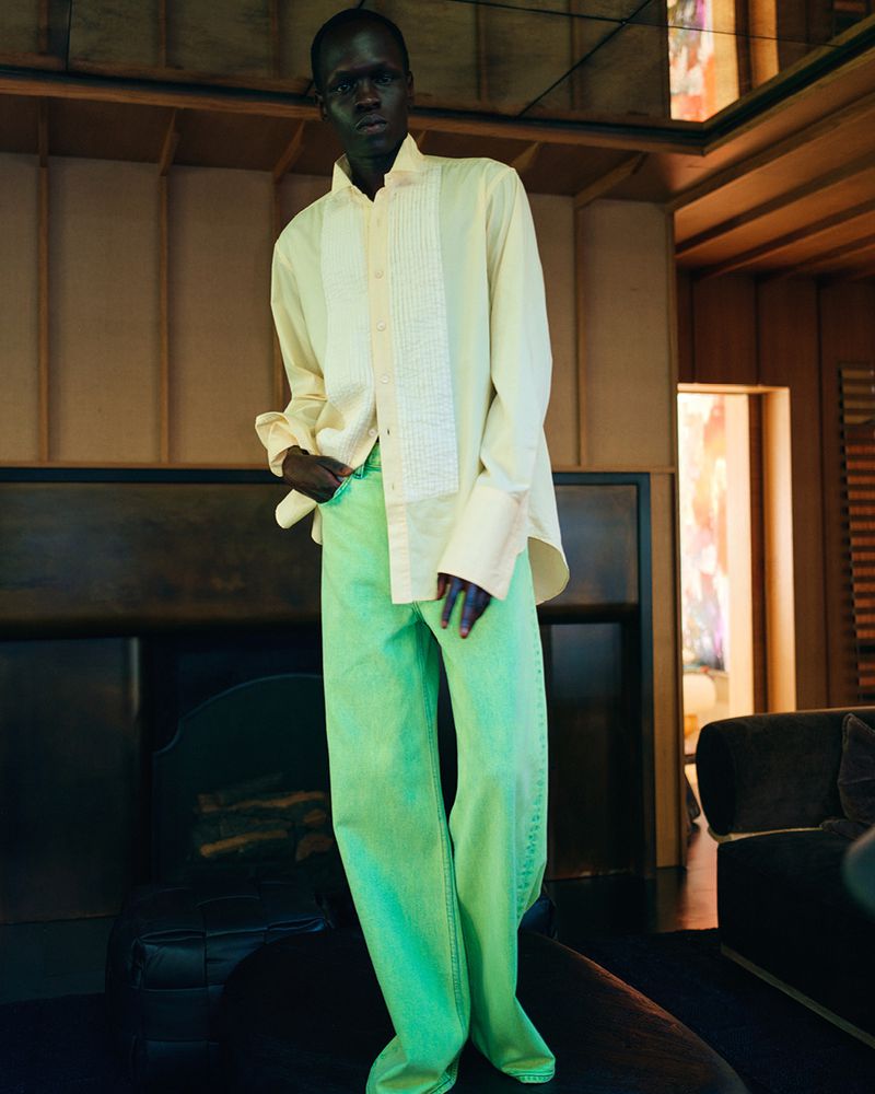 Male model posing in a pale yellow shirt and light green pants in an indoor setting with a fireplace, looking directly at the camera.