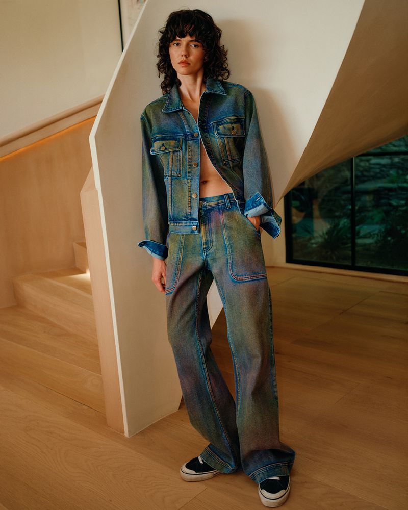 A young woman poses indoors wearing a blue and green tie-dye denim jacket and matching wide-leg jeans and black sneakers.