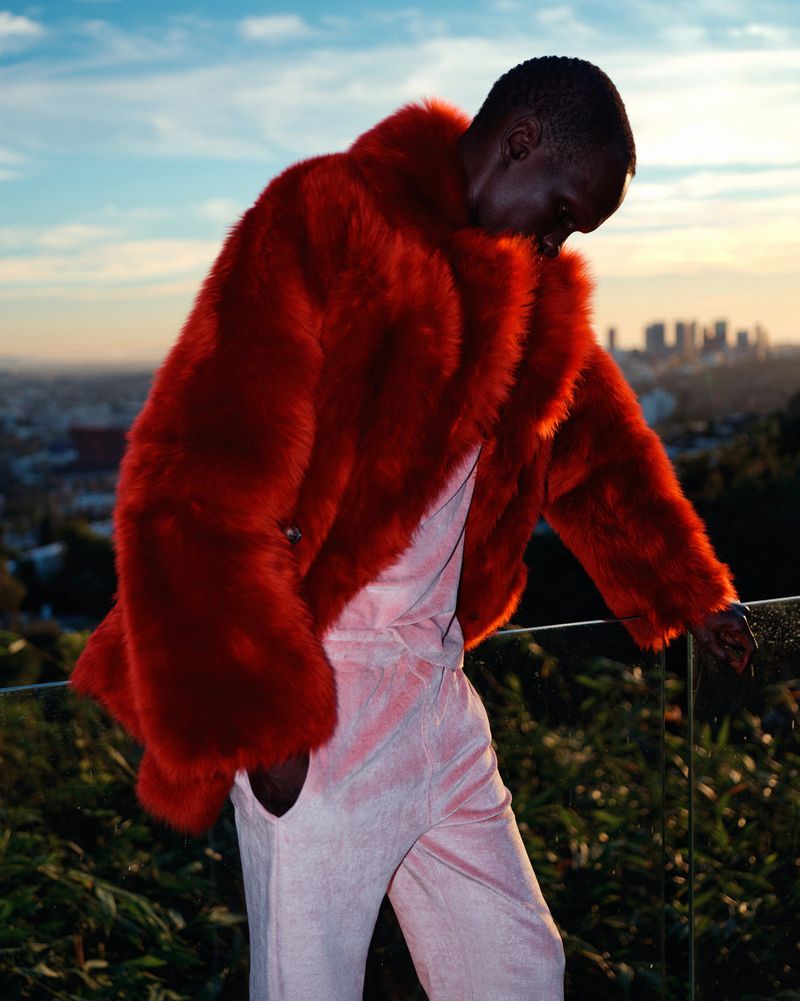 An adult male wearing a red fur coat and pink suit stands outside with a city skyline, blue sky, and greenery in the background.
