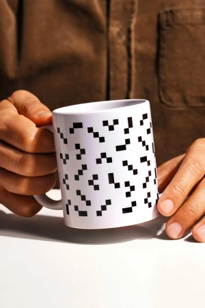 Close-up of a person holding a white coffee mug with a black pixel pattern against a neutral background in a studio setting.