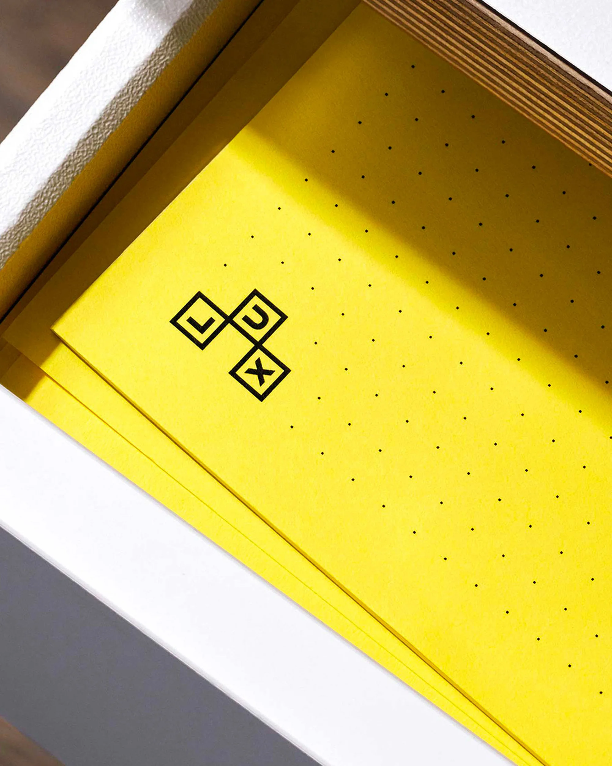 Yellow sheets of paper with a pattern of black dots and a black logo inside a white container, creating a minimalist and graphic composition.