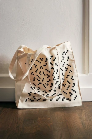 Beige tote bag with black pixelated pattern resting against a white wall and dark wood floor. The bag has two handles.