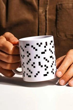 Person holding a white ceramic coffee mug patterned with small black pixelated blocks against a plain white surface