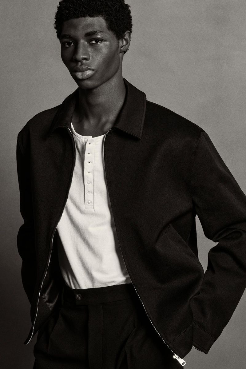 Portrait of a Black male model in a black jacket and pants against a grey backdrop, looking at the camera with neutral expression.