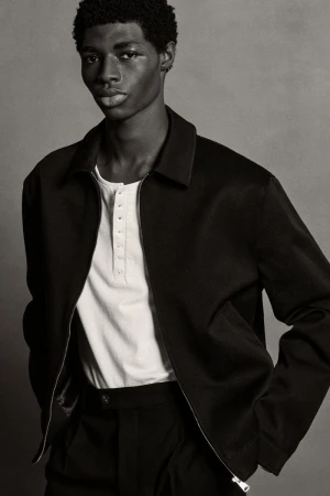 Portrait of a Black male model in a black jacket and pants against a grey backdrop, looking at the camera with neutral expression.