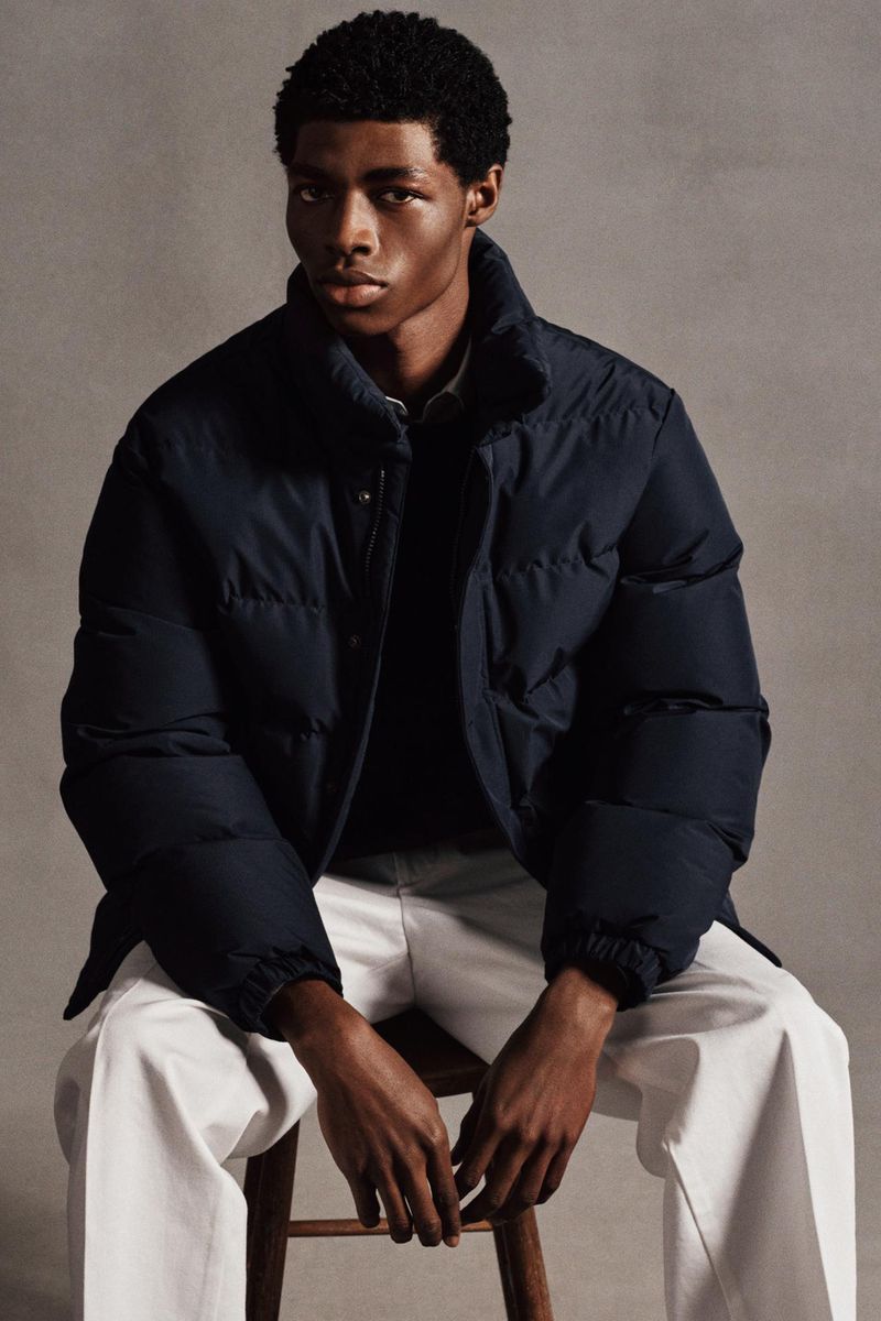 A stylish dark-skinned male model in a navy puffer jacket sits on a wooden stool, set against a plain grey backdrop in a studio setting.