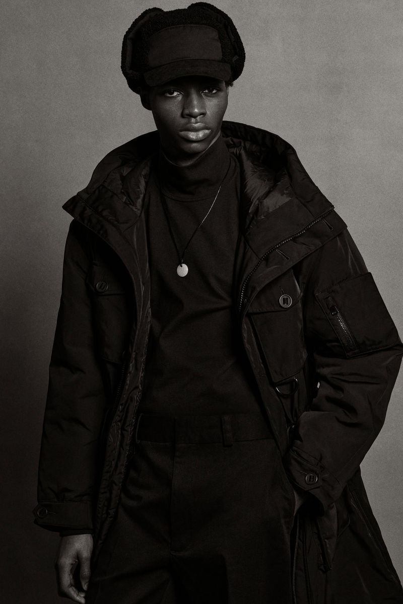 Black male model in black fur hat and black coat stands in a photo studio with a gray backdrop. Full fashion shot.