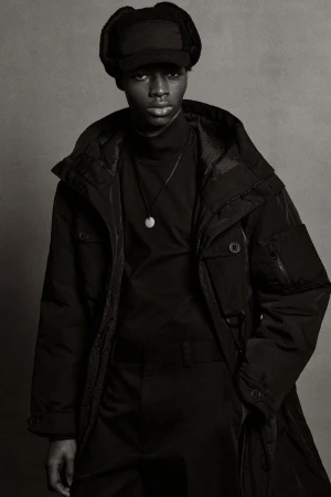 Black male model in black fur hat and black coat stands in a photo studio with a gray backdrop. Full fashion shot.