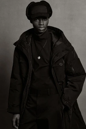 Portrait of a black male model in a black winter coat and trapper hat against a neutral textured background