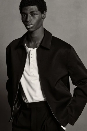 Black man wearing dark zip-up jacket and white shirt against a neutral grey studio background