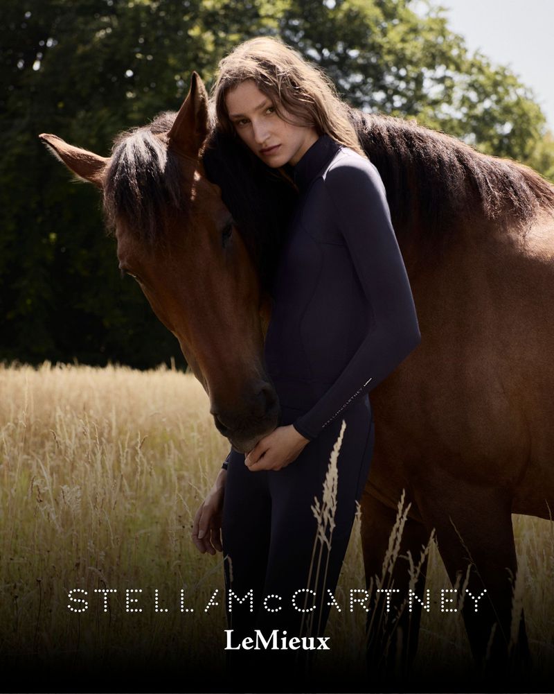 Woman in athletic wear stands next to a brown horse in a field with tall grasses, with dappled sunlight and STELLAMCCARTNEY and LeMieux logos.