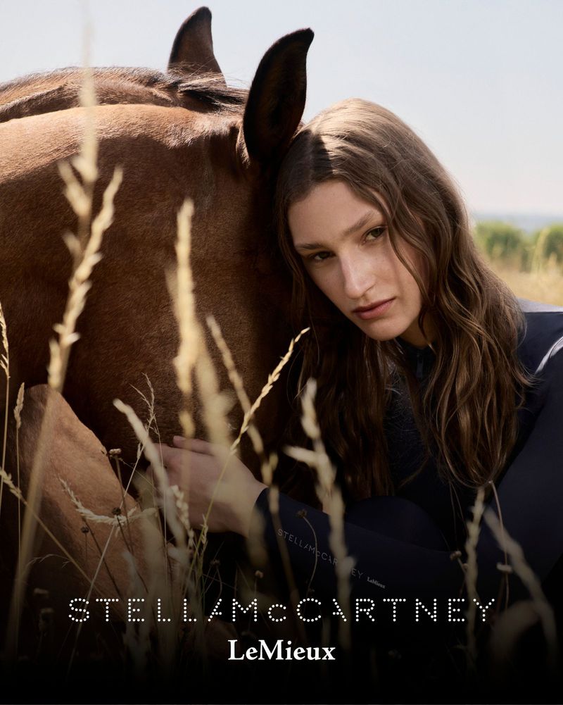 Portrait of a woman with long hair, leaning against a brown horse outdoors with tall grasses, under a bright sky in soft, natural light.