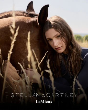 Portrait of a woman with long hair, leaning against a brown horse outdoors with tall grasses, under a bright sky in soft, natural light.