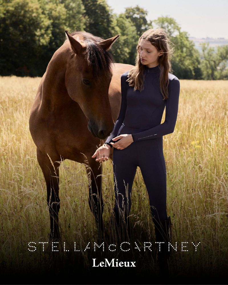 Woman in a dark blue jumpsuit stands with a brown horse in a field with tall grasses, trees and sky in the background.