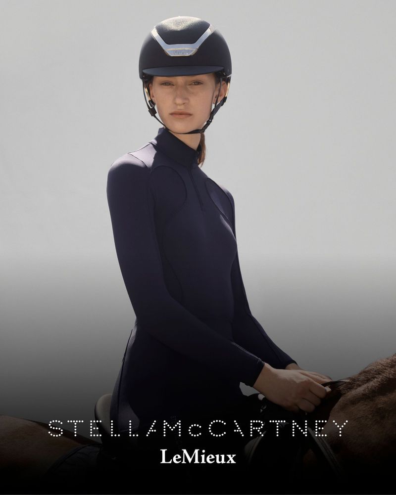 Woman in equestrian helmet and long-sleeved shirt, seated on a horse, facing forward against a gradient grey background.