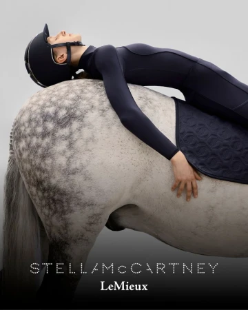 Woman in riding attire reclines on a light gray horse with a dark saddle pad against a pale background in a fashion advertisement.