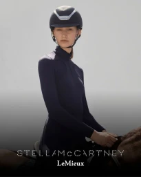 Model wearing a dark equestrian helmet and navy riding jersey seated on a horse against a pale gradient background.