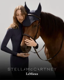 Model in navy equestrian top embracing a brown horse wearing a dark ear bonnet against a gradient grey studio backdrop.