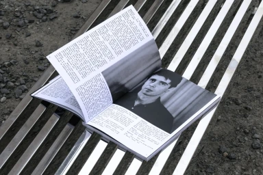 An open magazine lies on a metallic slatted bench, showing printed text and a black and white portrait photograph.