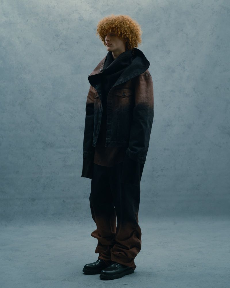 A stylish adult male with curly hair poses in a studio, wearing a dark, oversized denim jacket, black top, and brown-black gradient pants with black boots.