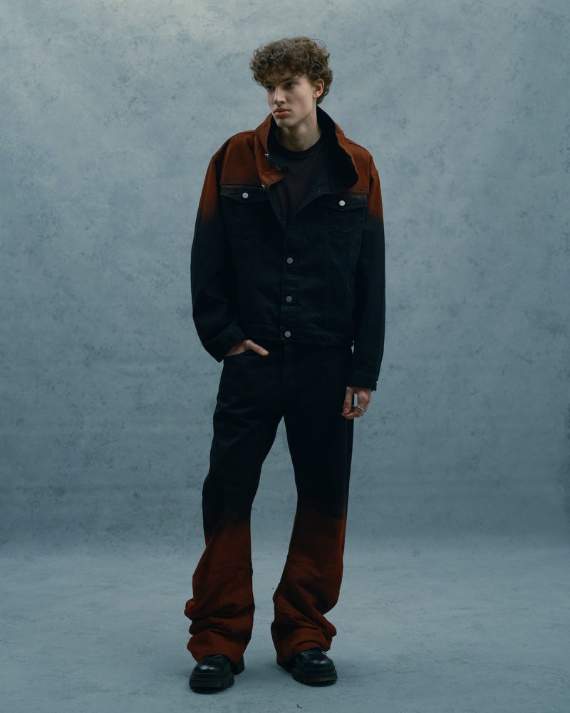 Male model stands in studio wearing black and red ombre denim jacket and matching oversized jeans, styled with black boots.