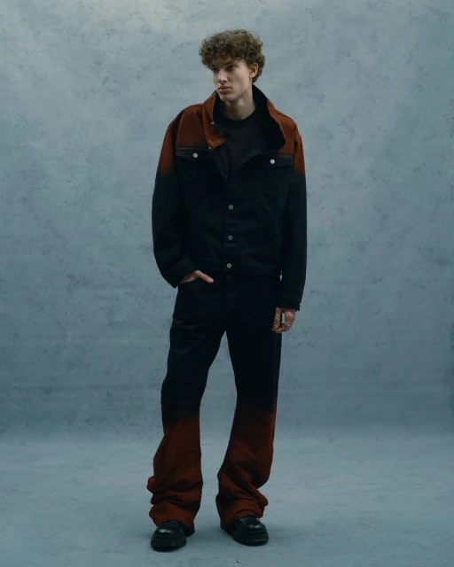 Male model stands in studio wearing black and red ombre denim jacket and matching oversized jeans, styled with black boots.