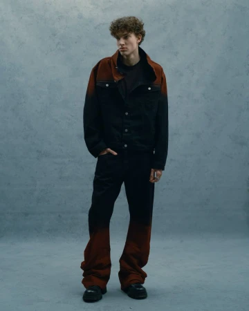 Male model stands in studio wearing black and red ombre denim jacket and matching oversized jeans, styled with black boots.