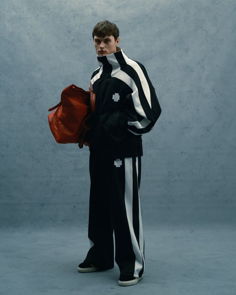 A young adult man stands in a light gray studio wearing a black and white track suit while holding a red leather duffel bag.