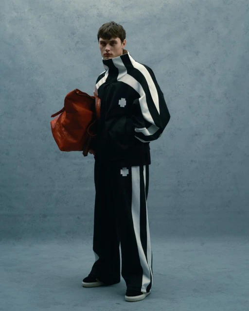 A young adult man stands in a light gray studio wearing a black and white track suit while holding a red leather duffel bag.