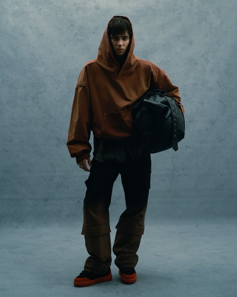 A person wearing a brown hooded sweatshirt, black pants, and orange sneakers, holding a black bag against a textured blue background.