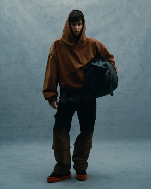 A person wearing a brown hooded sweatshirt, black pants, and orange sneakers, holding a black bag against a textured blue background.